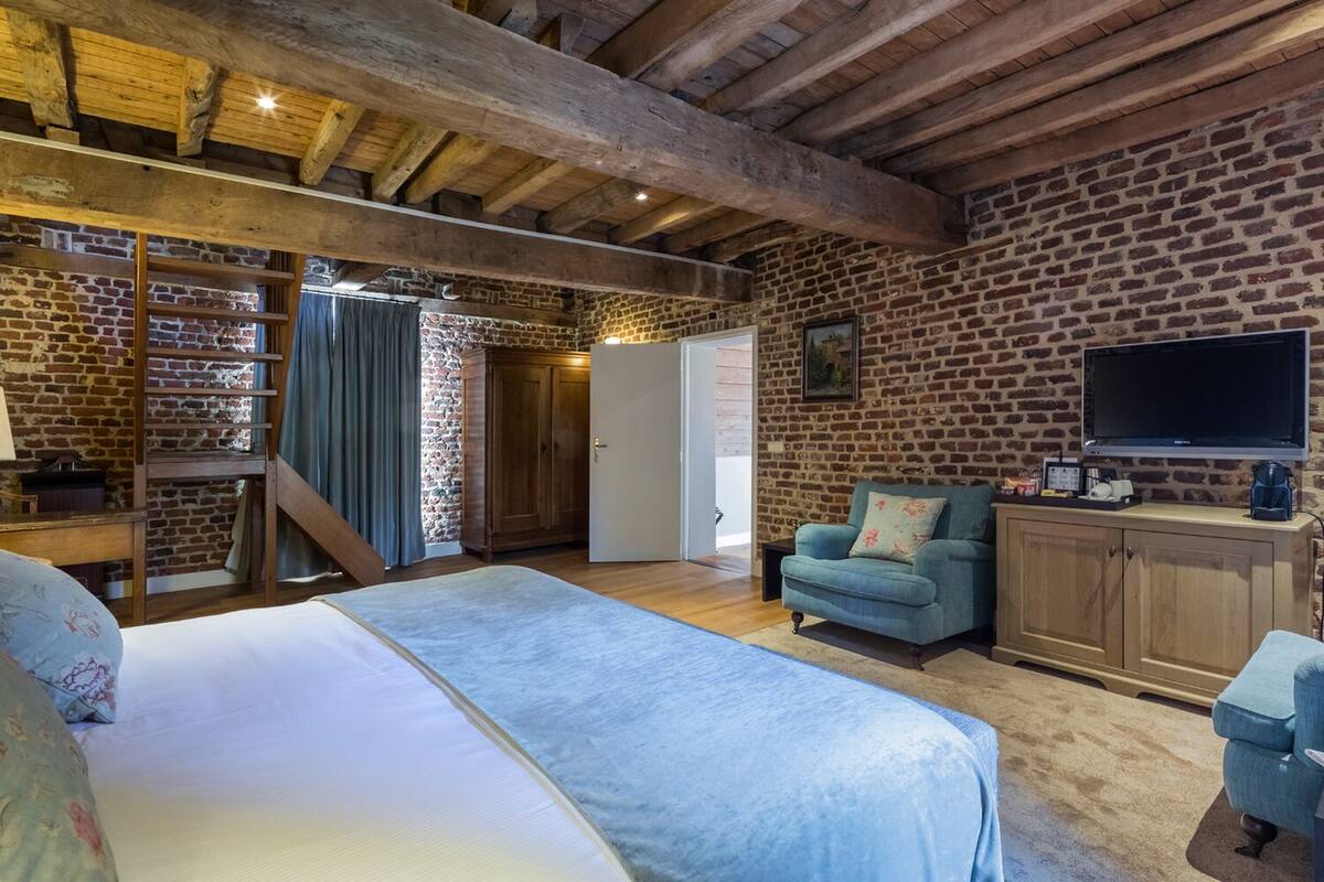 A bedroom with a large bed, a wooden ceiling, brick walls, a TV, and a cabinet.