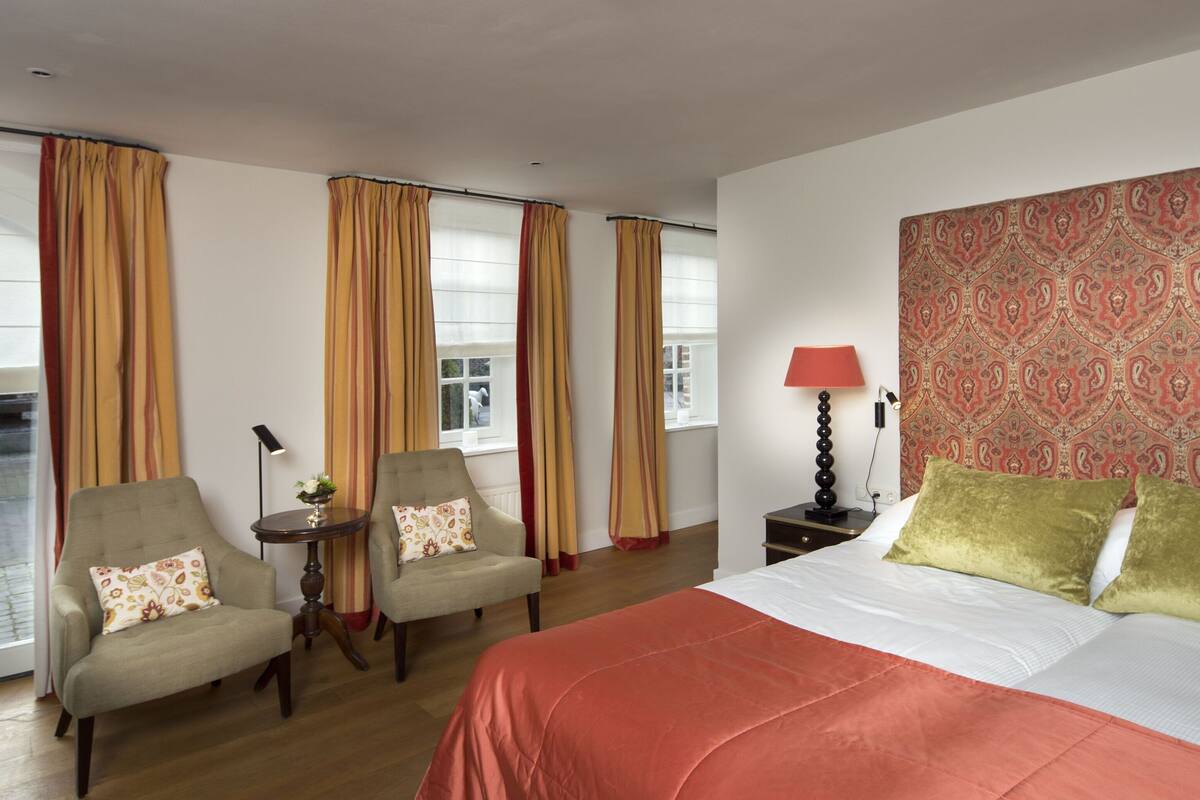 A hotel room with a large bed, two armchairs, a small table with a lamp, and a patterned wall.