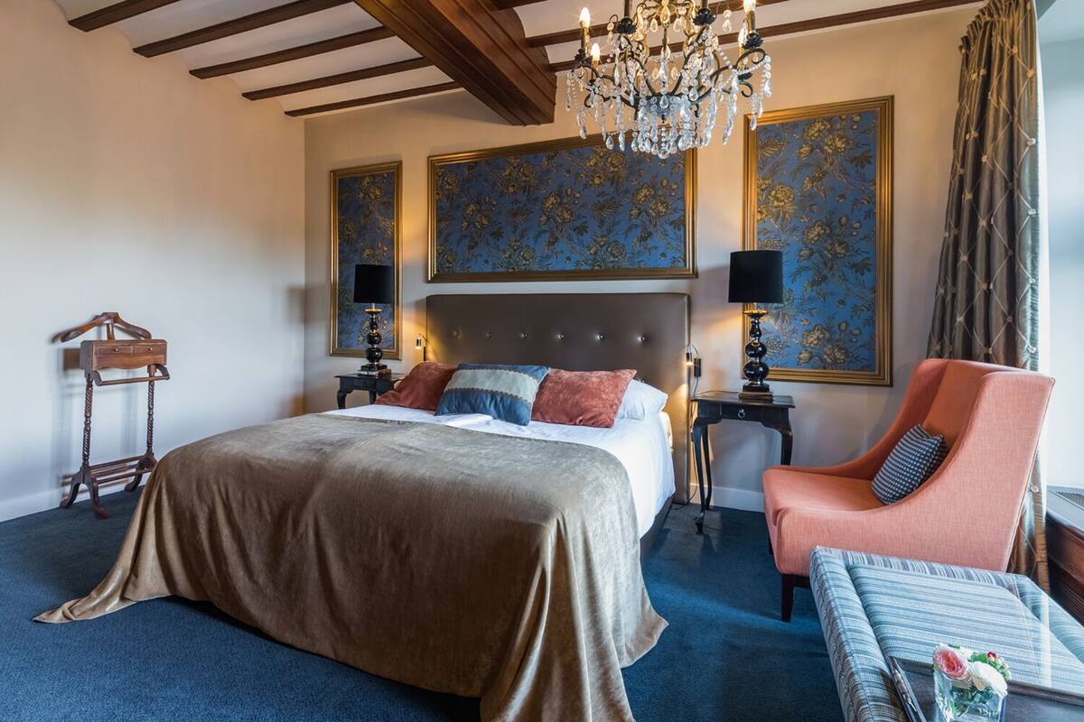 A hotel room with a large bed, a chandelier, a chair, and a table with a vase.