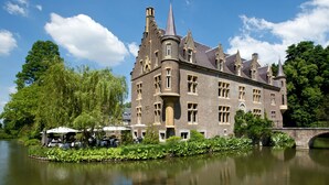 A historic building with a tower, a courtyard with tables and umbrellas, and a bridge over a calm river. - Van der Valk Hotel Kasteel Terworm (Heerlen)