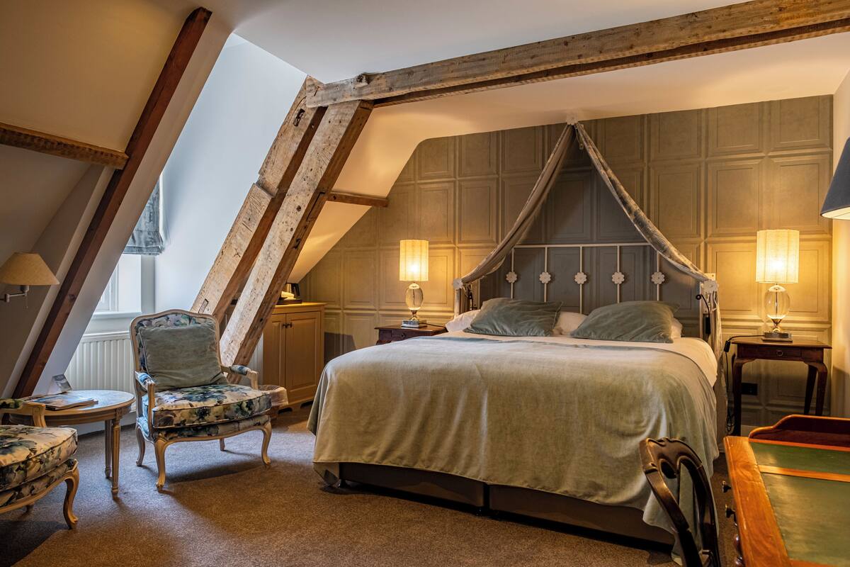 A bedroom with a sloped ceiling, a four-poster bed, two bedside lamps, a chair, and a small table.