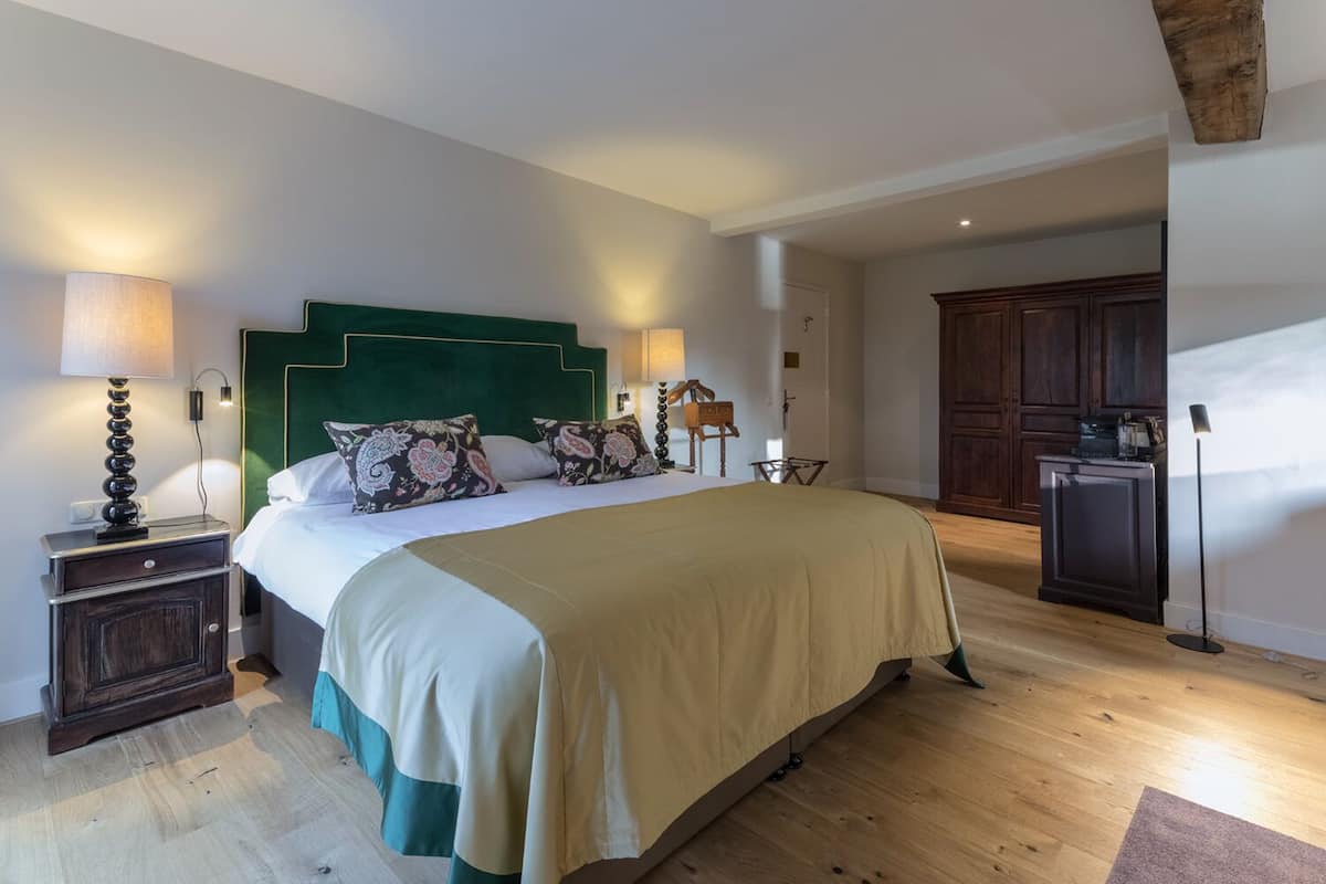 A bedroom with a large bed, a nightstand, a dresser, and a desk with a lamp.
