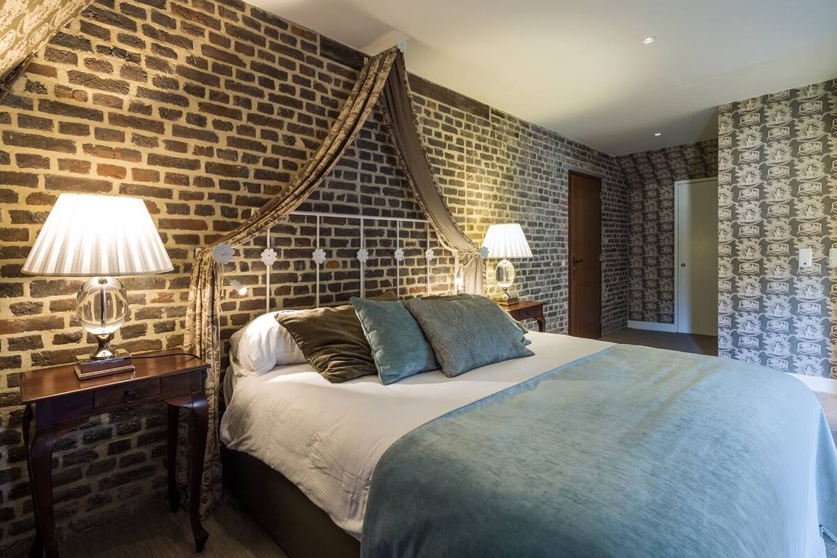 A bedroom with a brick wall, a canopy bed, two bedside lamps, and a patterned wallpaper on the adjacent wall.