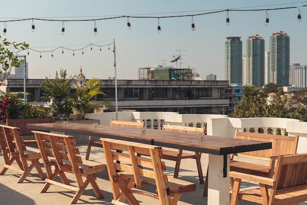 An outdoor seating area with wooden tables and chairs, string lights, and a view of tall buildings in the background.
