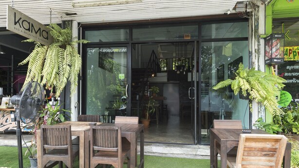 An outdoor seating area with wooden tables and chairs, a large fern plant, and a sign for "KAMA BED & BREAKFAST BANGKOK". - Kama Bangkok - Boutique Bed & Breakfast - Hostel (Bangkok)