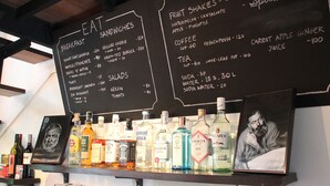 A menu board with various food and drink options, a shelf with liquor bottles, and a framed picture. - Kama Bangkok - Boutique Bed & Breakfast - Hostel (Bangkok)