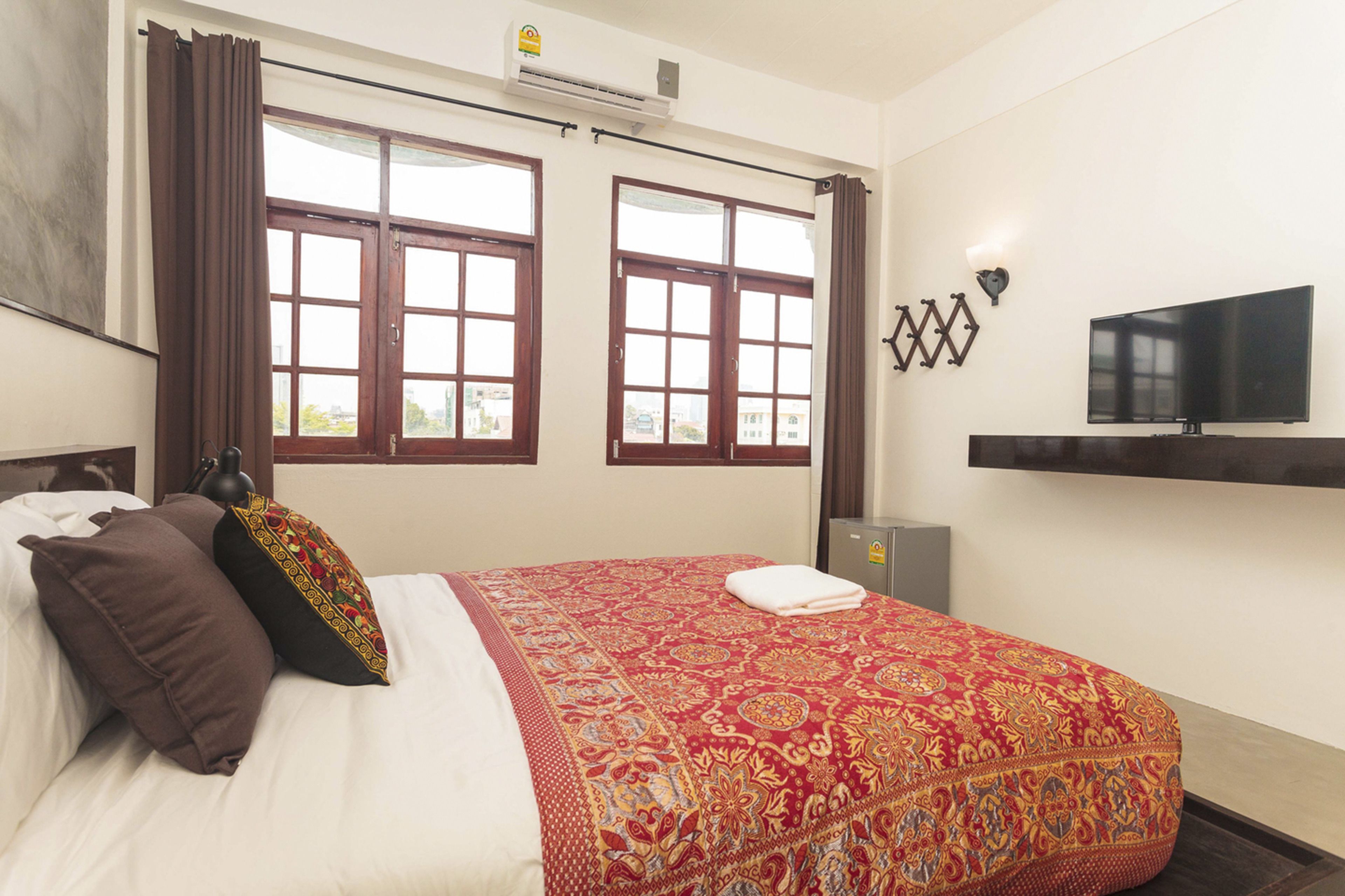 A hotel room with a large bed, two windows with curtains, a flat-screen TV, and a wall-mounted shelf.