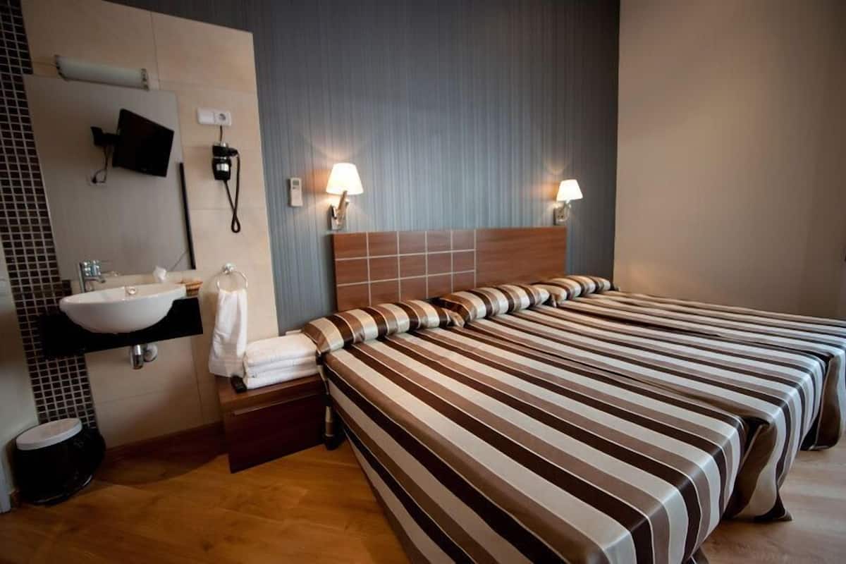 Habitación de hotel con una cama grande, una mesita de noche, un televisor montado en la pared, un baño con lavamanos y espejo, y dos lámparas de pared.