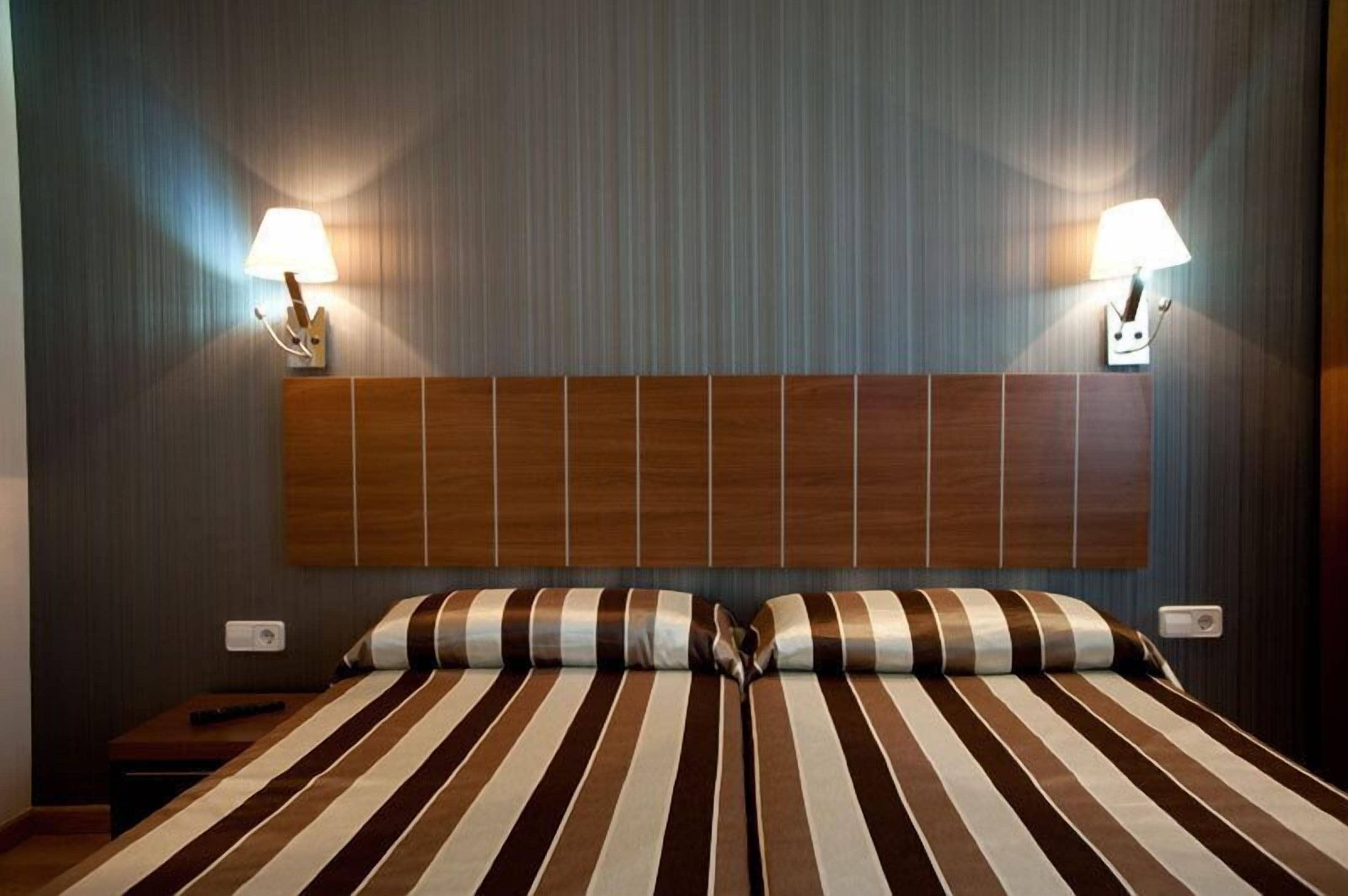 Une chambre d’hôtel avec un lit à rayures, une tête de lit en bois, deux lampes fixées au mur et une table de chevet avec une télécommande.