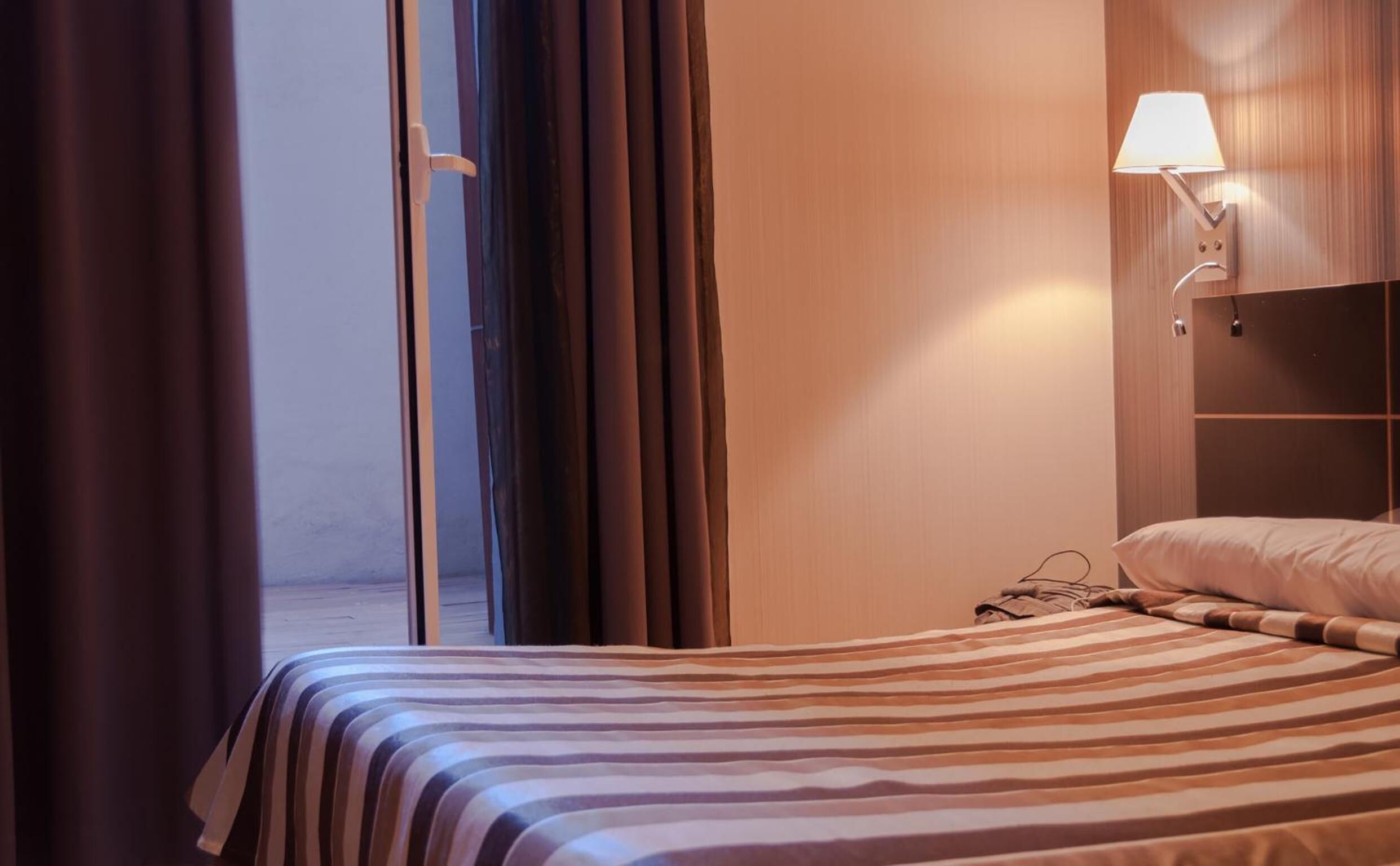 A hotel room with a bed, a nightstand, a lamp, and a view of the outside.