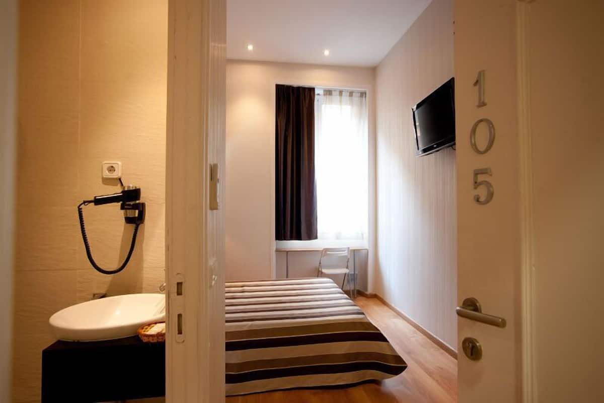 Habitación de hotel con cama, un escritorio pequeño, ventana con cortinas, televisor montado en la pared y baño con inodoro y lavamanos.