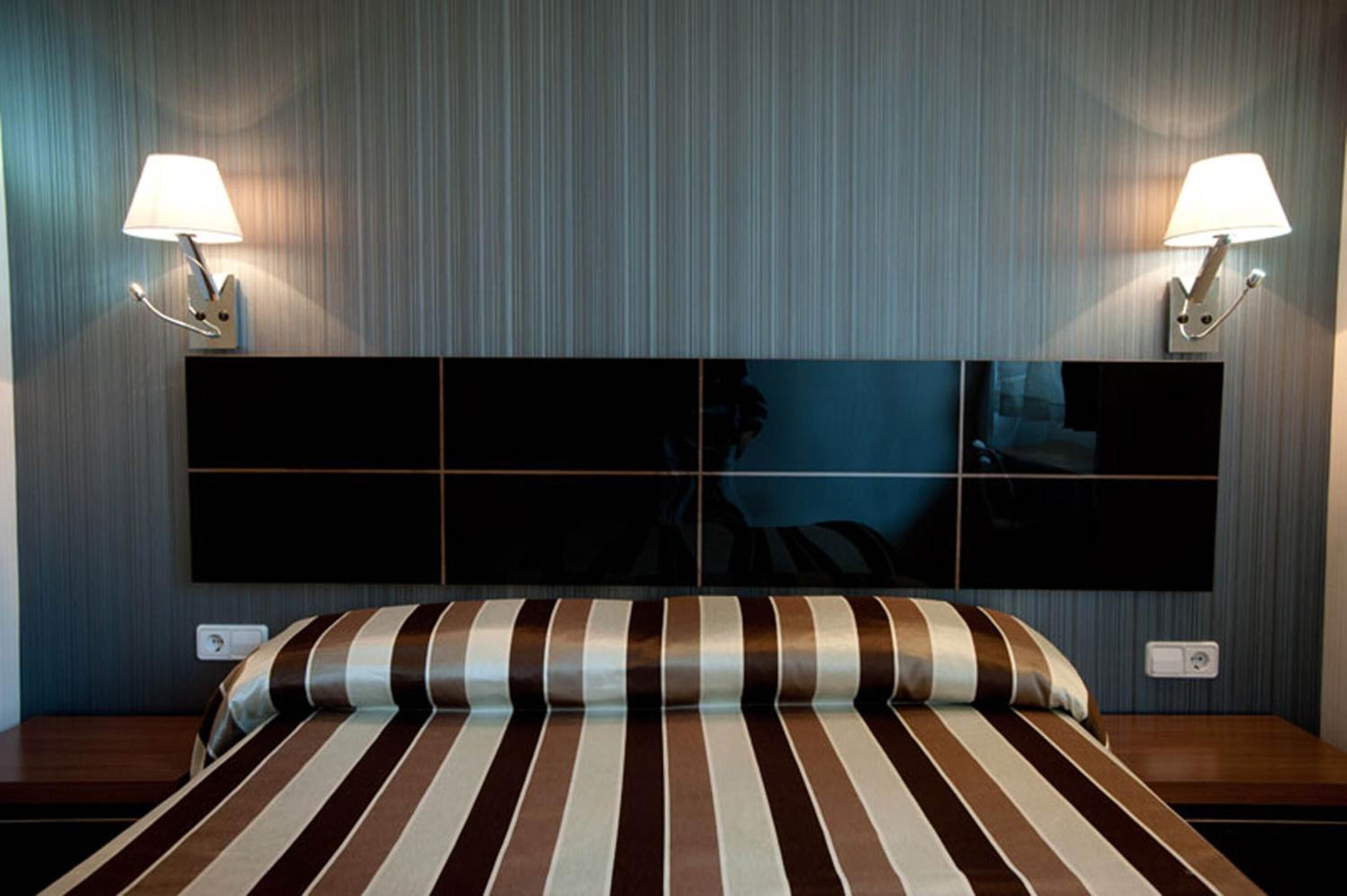 A hotel room with a large bed, striped bedding, two wall-mounted lamps, and a large mirror above the bed.