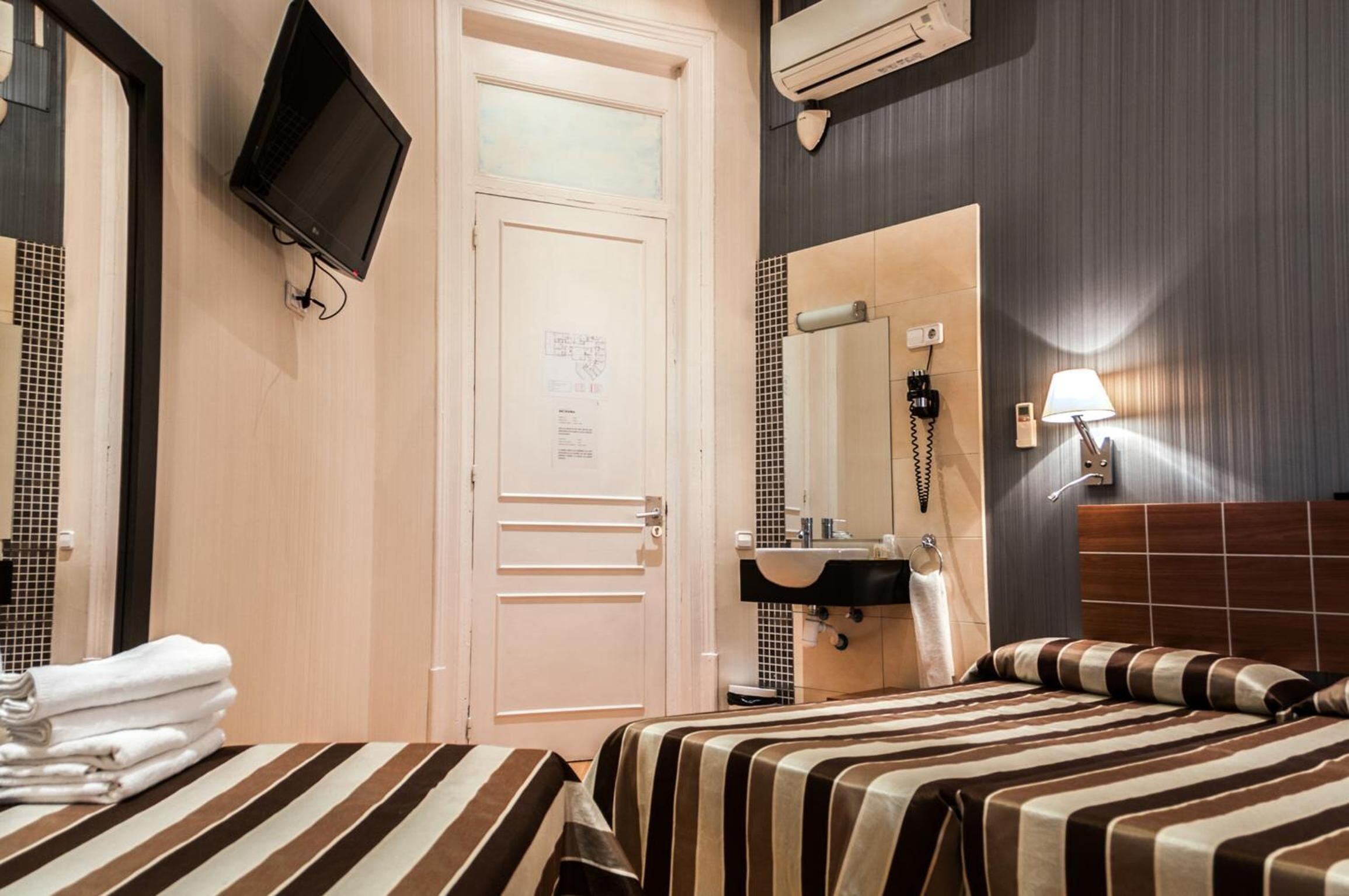 A hotel room with a bed, a bedside table with towels, a television mounted on the wall, a mirror, a sink, and a telephone.