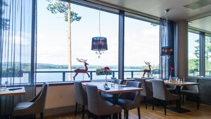 Un restaurante con grandes ventanales que dan a una masa de agua, con mesas y sillas de madera, estatuas decorativas de ciervos y lámparas colgantes.