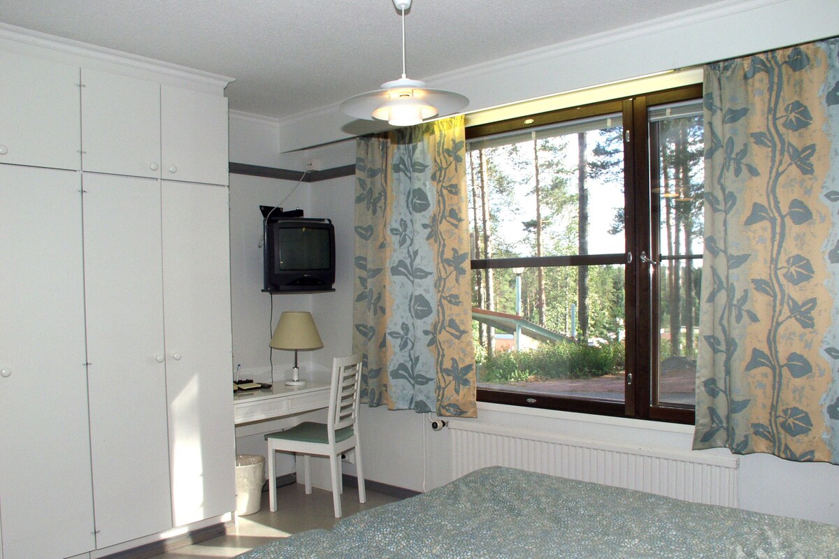 A bedroom with a bed, a desk with a chair, a television, and a window with curtains.