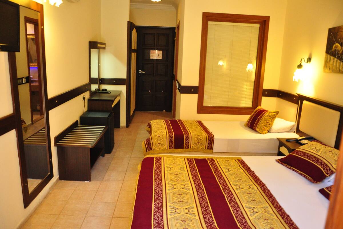 Une chambre d’hôtel avec deux lits, un banc, un bureau, un miroir et une fenêtre avec des stores.