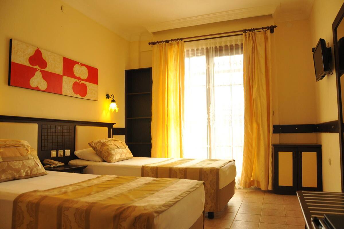 Une chambre d’hôtel avec deux lits, un téléviseur fixé au mur et une fenêtre avec des rideaux jaunes.