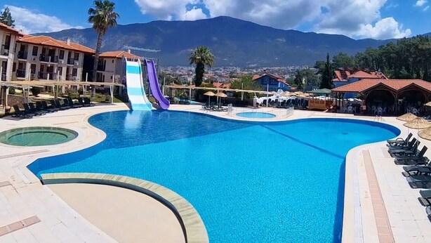 A large swimming pool with a slide, surrounded by lounge chairs and umbrellas, with a mountain view in the background. - Telmessos Neva Hotel - Halal-Non Alcoholic  Hotel (Fethiye)