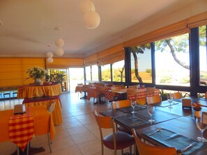 Un restaurante con varias mesas y sillas, ventanas amplias que dan a árboles y una carretera, y farolillos de papel colgantes.