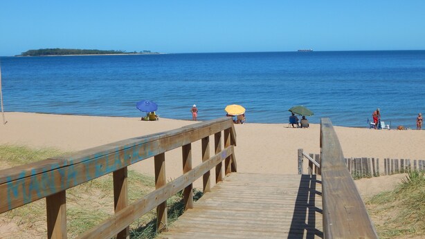Una pasarela de madera que conduce a una playa de arena con sombrillas y gente.