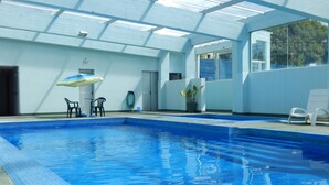 Una piscina interior con agua azul cristalina, techo de cristal y algunas sillas de descanso.