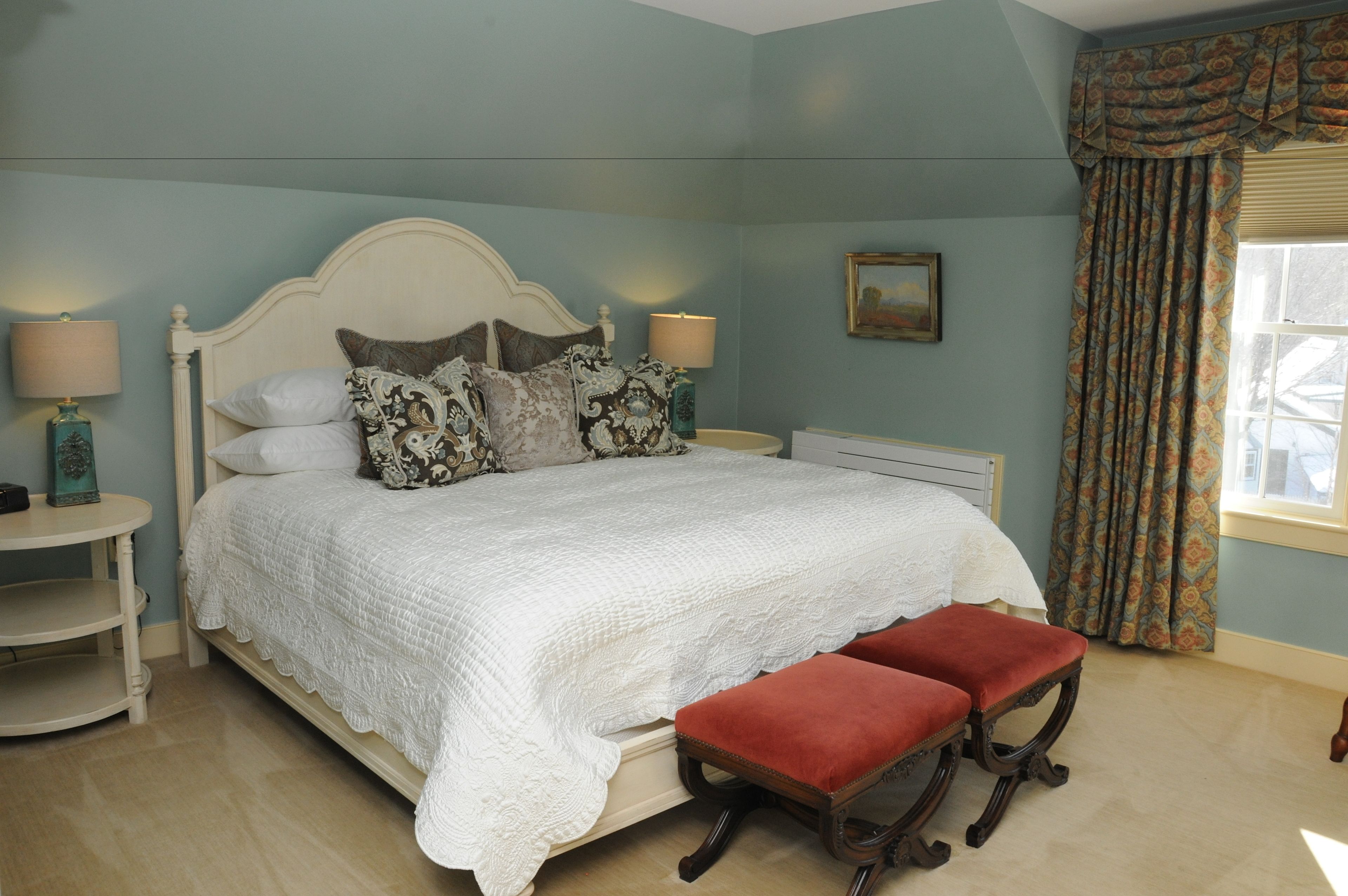 A bedroom with a large bed, two bedside tables, a red bench, and a window with curtains.