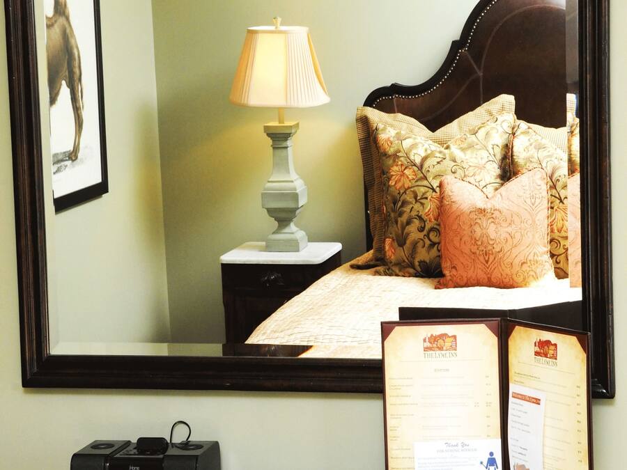 A hotel room with a large mirror, a bed with floral pillows, a bedside table with a lamp, a clock, and a menu.