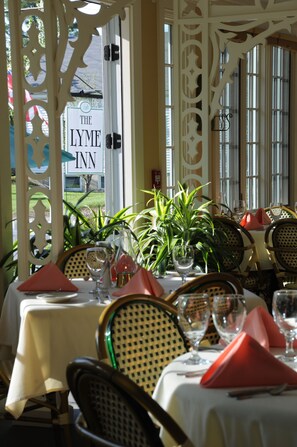 An indoor dining area with tables set for two, featuring white tablecloths, red napkins, and glassware.