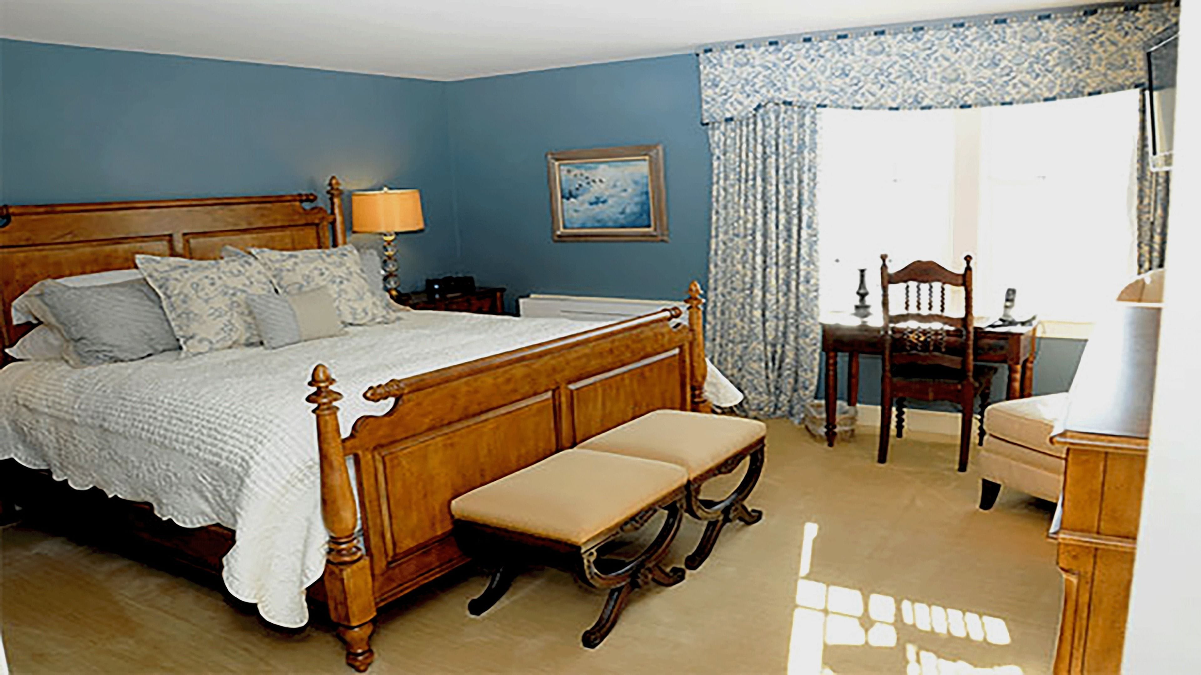 A bedroom with a wooden bed, a bench, a chair, a desk, a lamp, and a framed picture on the wall.