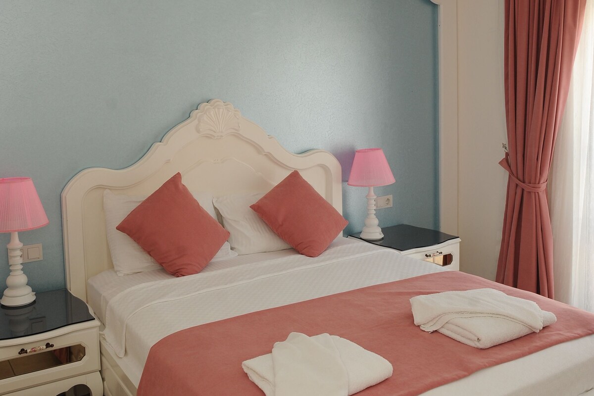 Una cama bien hecha con cabecera blanca, dos almohadas y colcha rosa. Hay dos mesitas de noche con lámparas, un ventilador de techo y una ventana con cortinas.