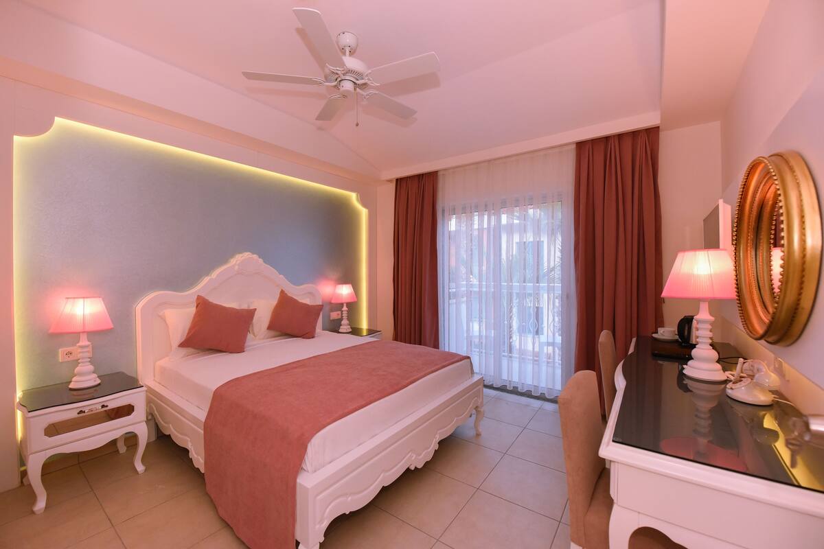 A bedroom with a bed, bedside tables, a desk, a chair, a ceiling fan, and a window with curtains.