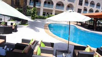 Zona de piscina con mobiliario de mimbre y sombrillas, piscina y un edificio con ventanas arqueadas y balcones.