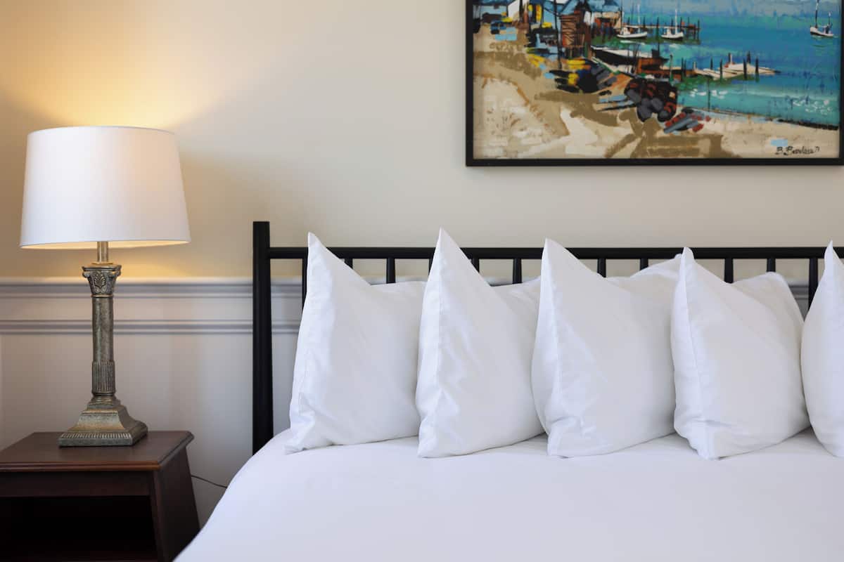 A bed with white pillows, a nightstand with a lamp, and a painting of a beach scene on the wall.