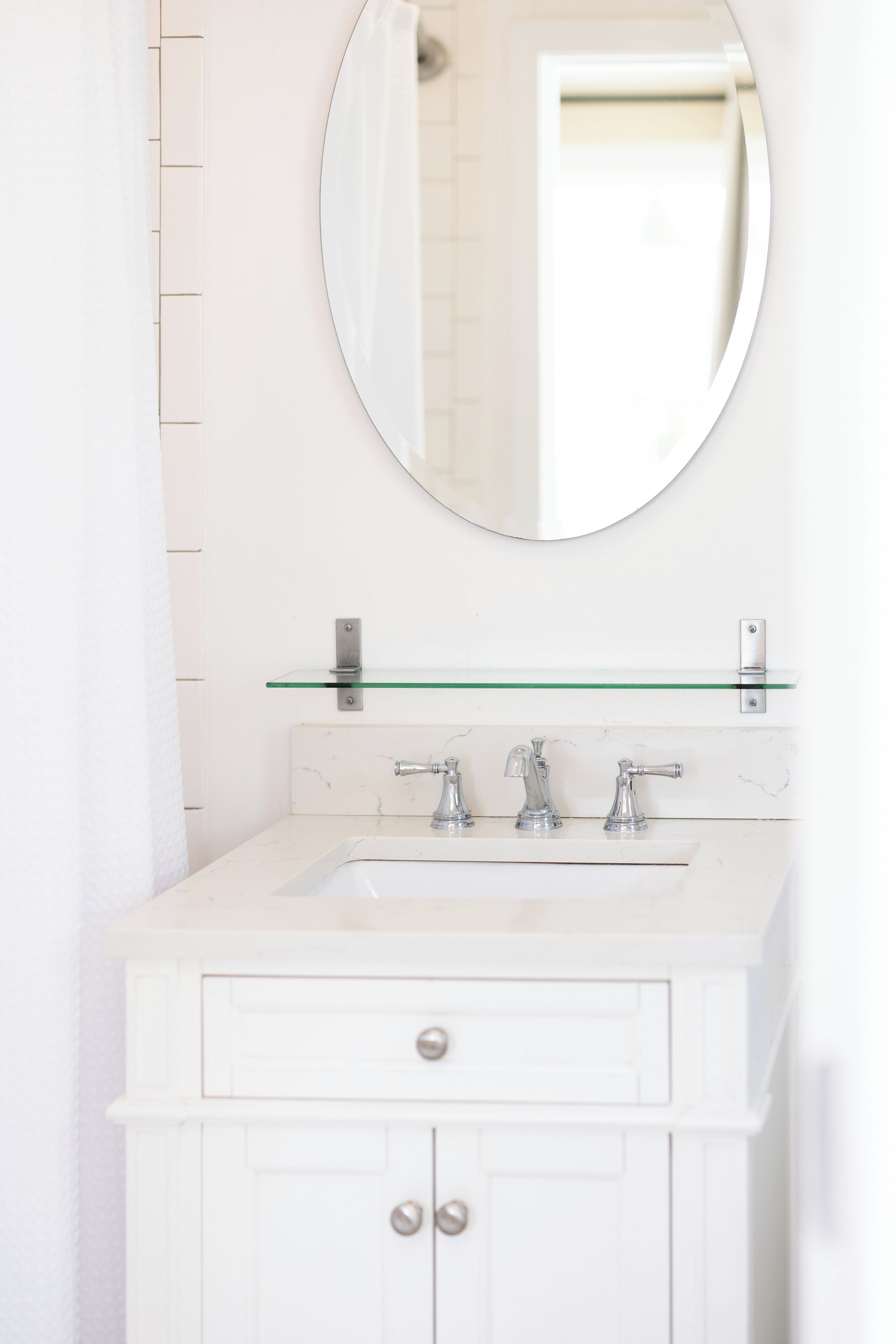 A bathroom with a white vanity, a round mirror, and a glass shelf.