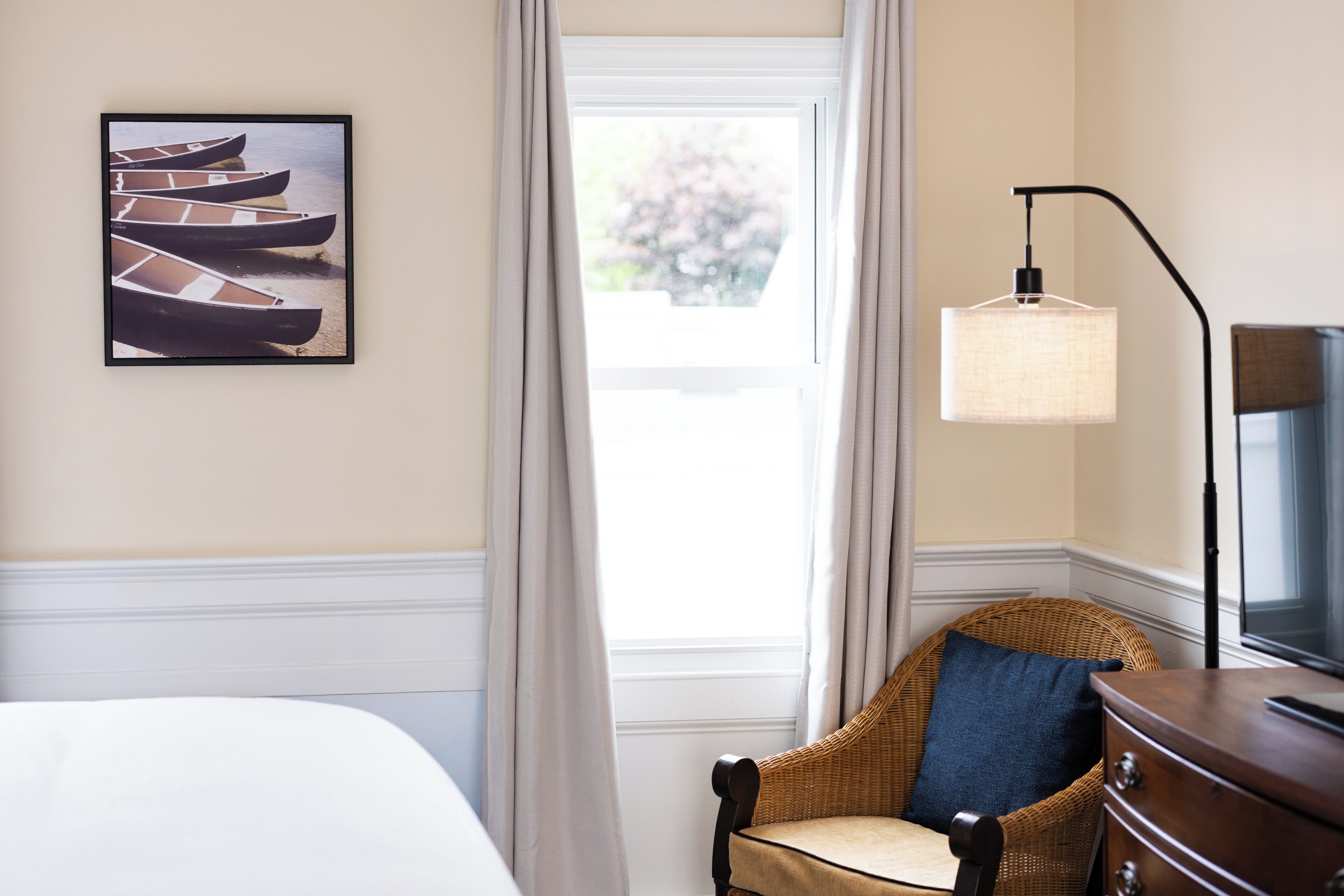 Una camera d'albergo con un letto, una sedia in vimini, una cassettiera, una lampada e un quadro appeso raffigurante barche.
