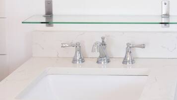 A bathroom with a white sink, two faucets, a glass shelf above the sink, and a round mirror.