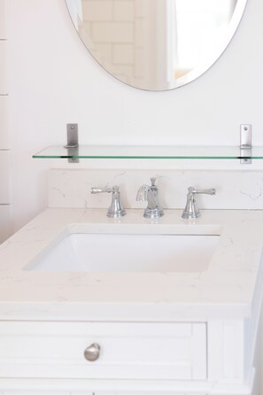 A bathroom with a white sink, two faucets, a glass shelf above the sink, and a round mirror. - Inn at 7 Central (Provincetown)