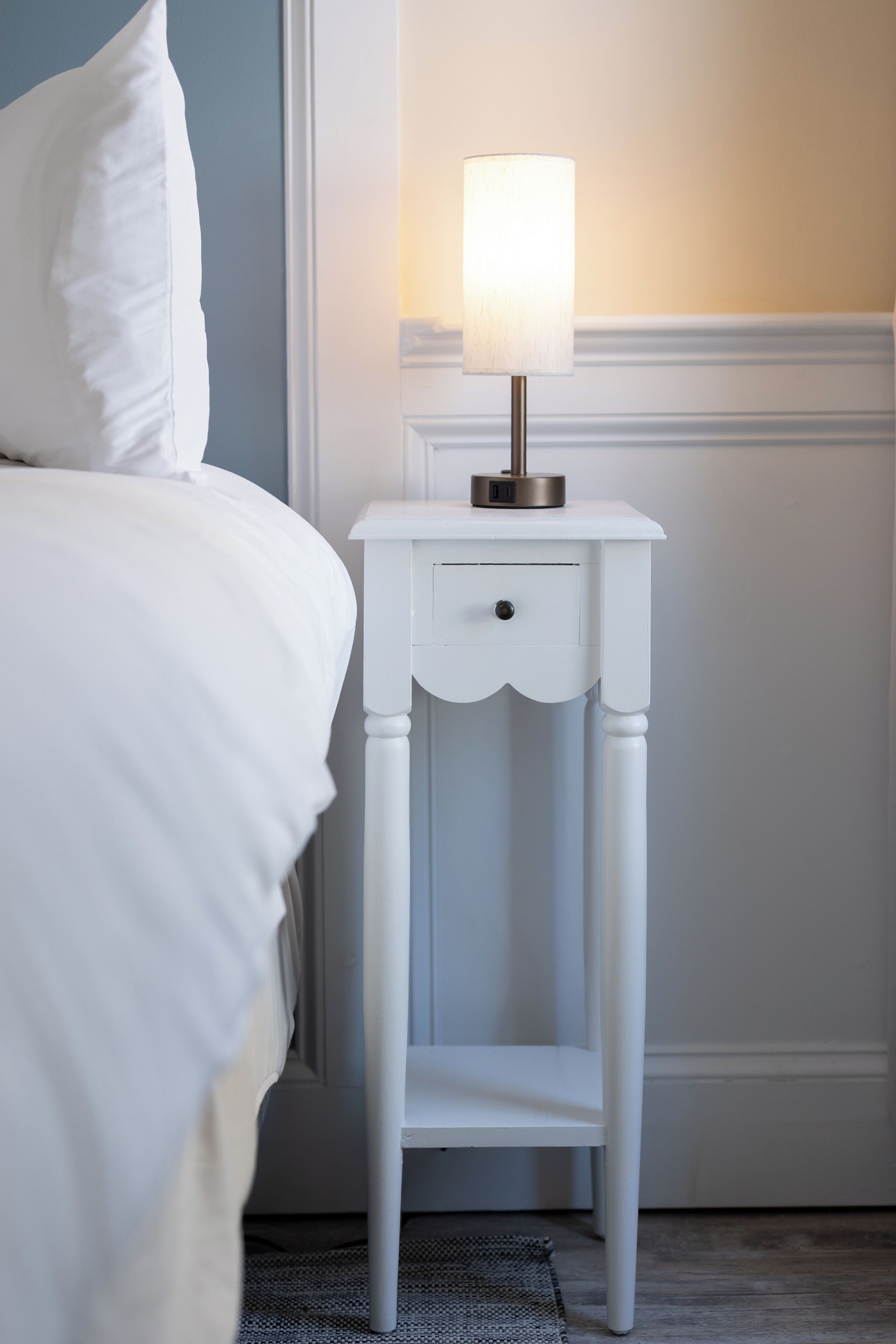 A white bedside table with a lamp, a bed with white bedding, and a plain wall.