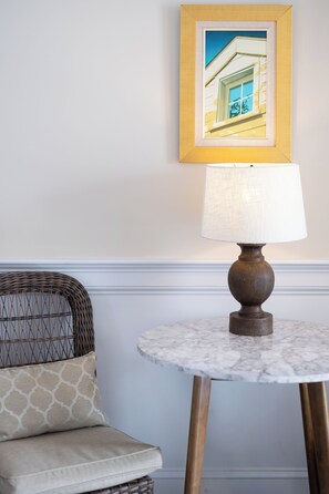 A wicker chair with a cushion, a round table with a lamp, and a framed picture on the wall. - Inn at 7 Central (Provincetown)