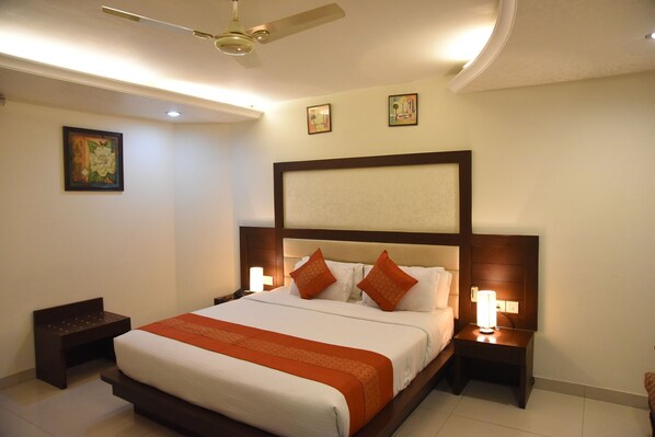 Quarto de hotel com uma cama grande, duas mesas de cabeceira com luminárias, um ventilador de teto e obras de arte nas paredes.