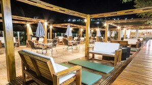 Een terras buiten met houten meubels, zoals banken en tafels, onder een houten pergola met lichtjes.