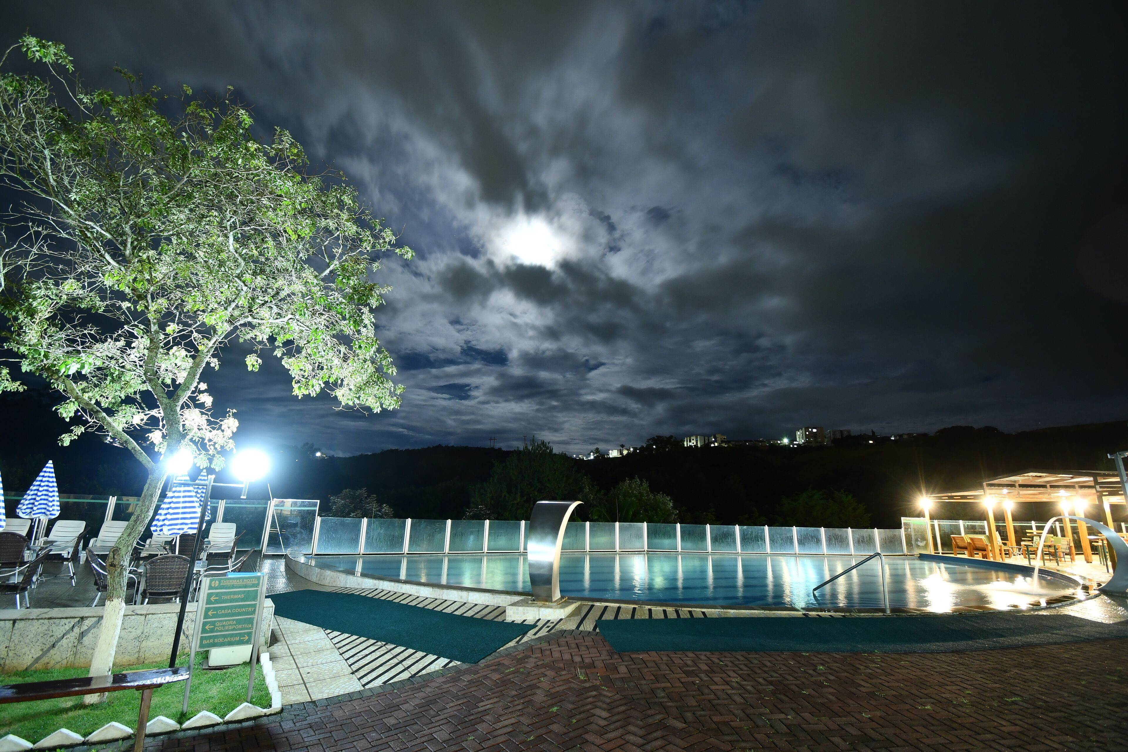 A swimming pool area with lounge chairs and umbrellas, surrounded by a glass fence, under a cloudy sky.