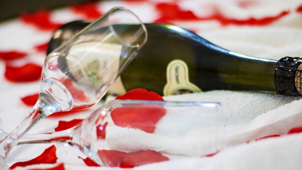 Une bouteille de champagne et un verre posés sur un lit, parsemé de pétales de roses rouges.