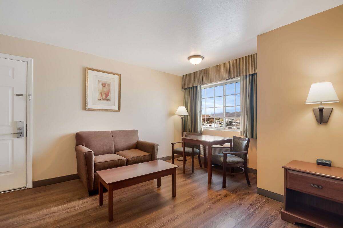 A hotel room with a sofa, a small table, a dining table with chairs, a lamp, and a window with curtains.