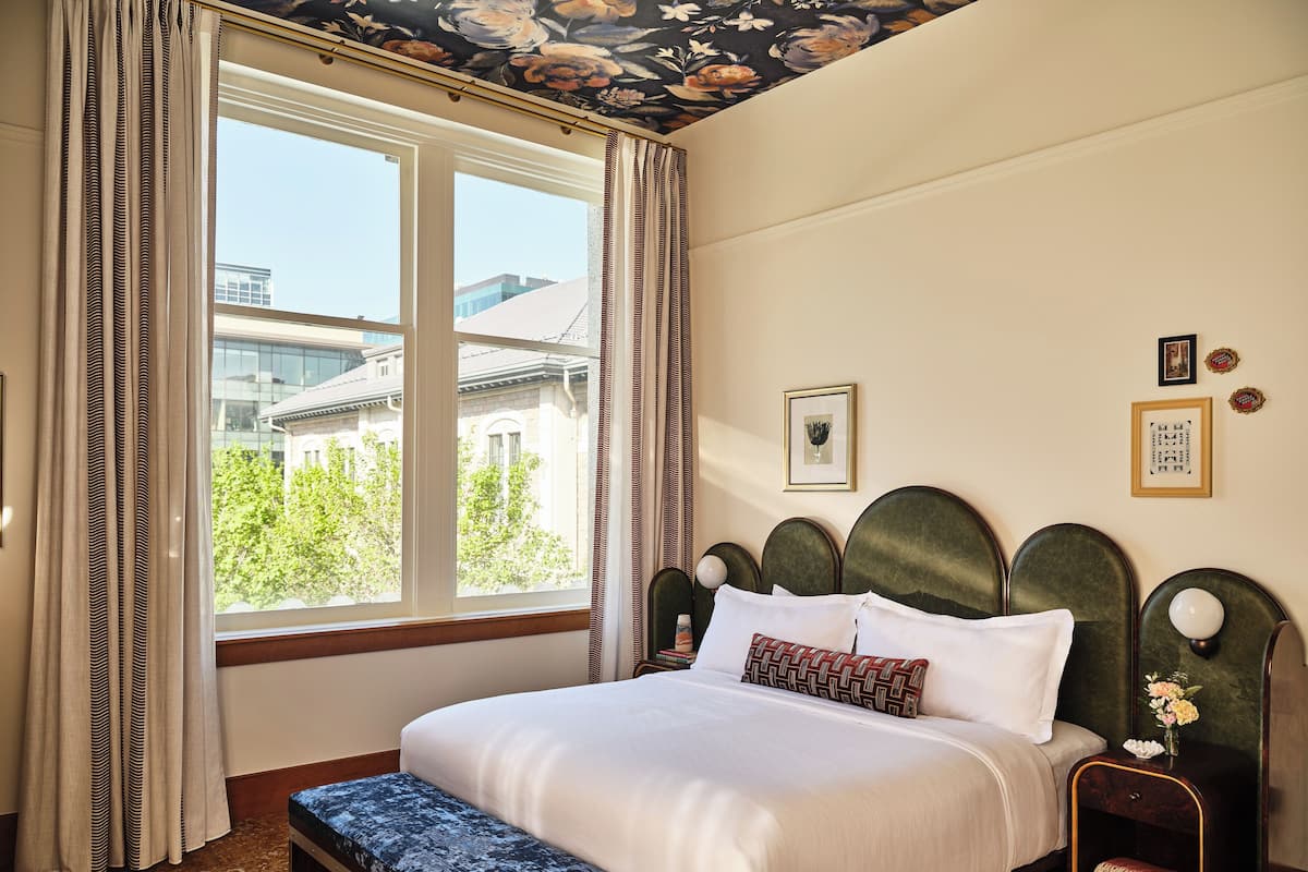 Une chambre d’hôtel dotée d’une grande fenêtre, d’un lit avec une tête de lit, d’un banc et d’une table de chevet avec une lampe.