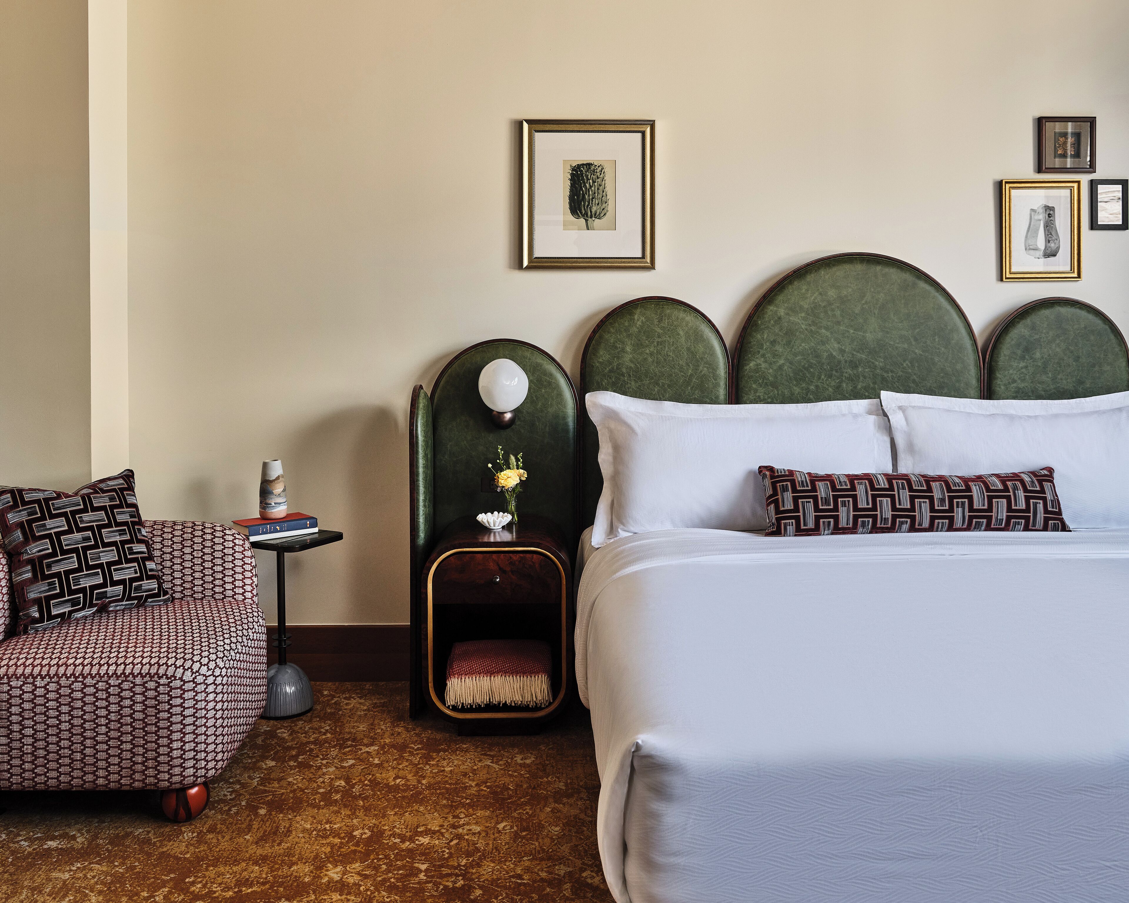 Une chambre d’hôtel avec un grand lit, une chaise, une table de chevet et une petite table. Des tableaux encadrés ornent le mur.