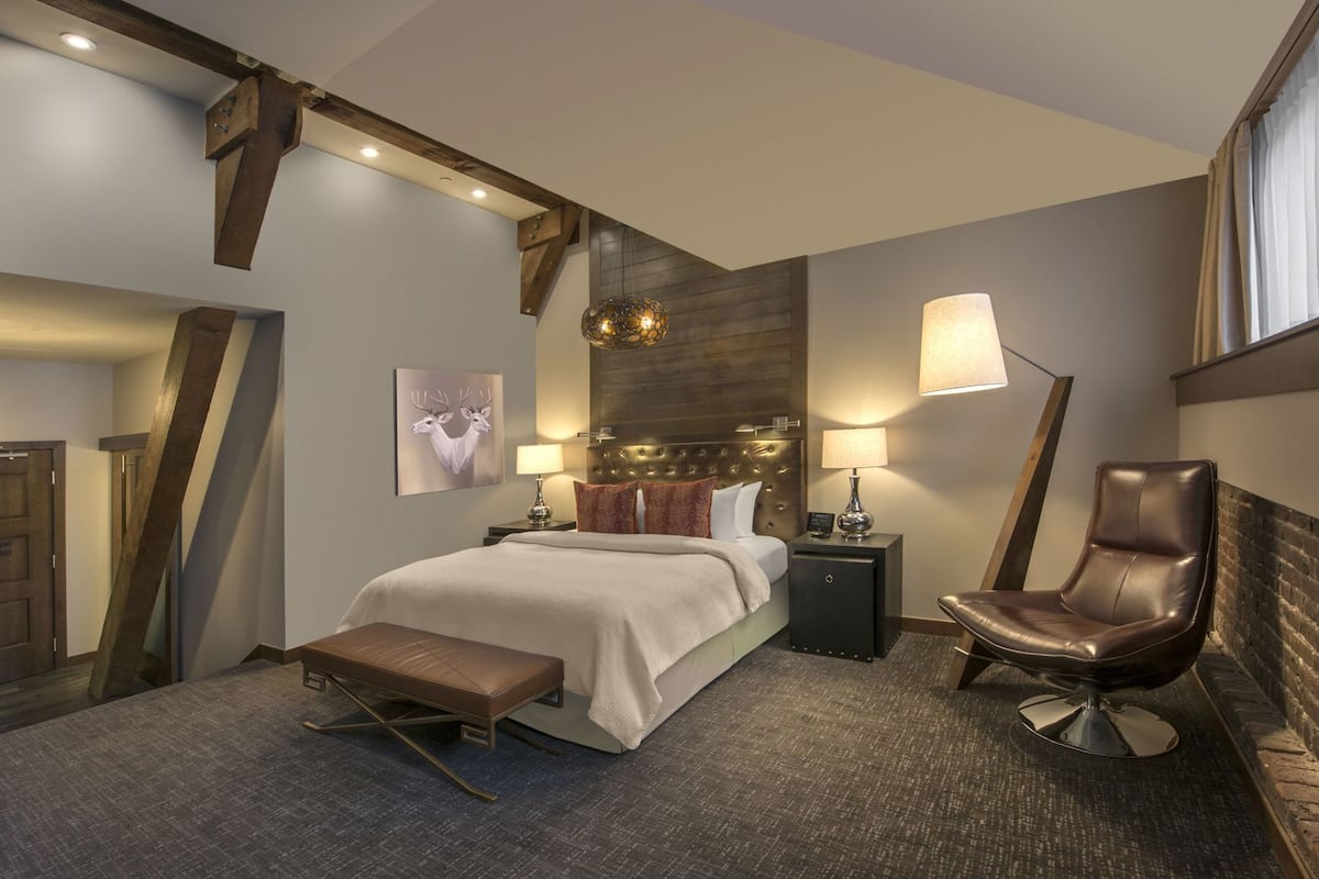 Une chambre d’hôtel moderne avec un grand lit, un banc, un fauteuil en cuir et une tête de lit ornée d’un panneau en bois.