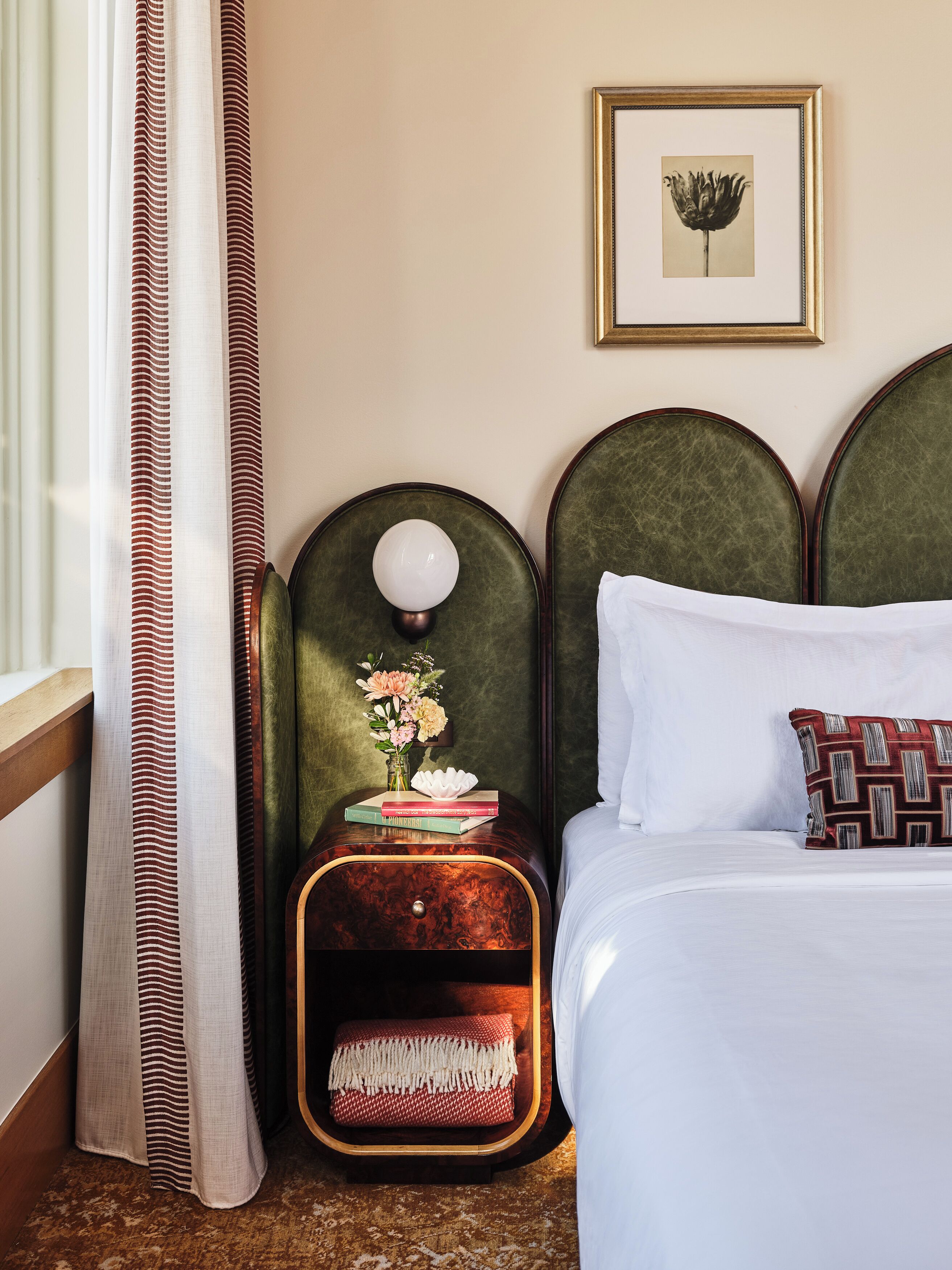 A neatly made bed with a white pillow and a patterned pillow, a bedside table with a lamp and a vase of flowers, and a framed picture on the wall.