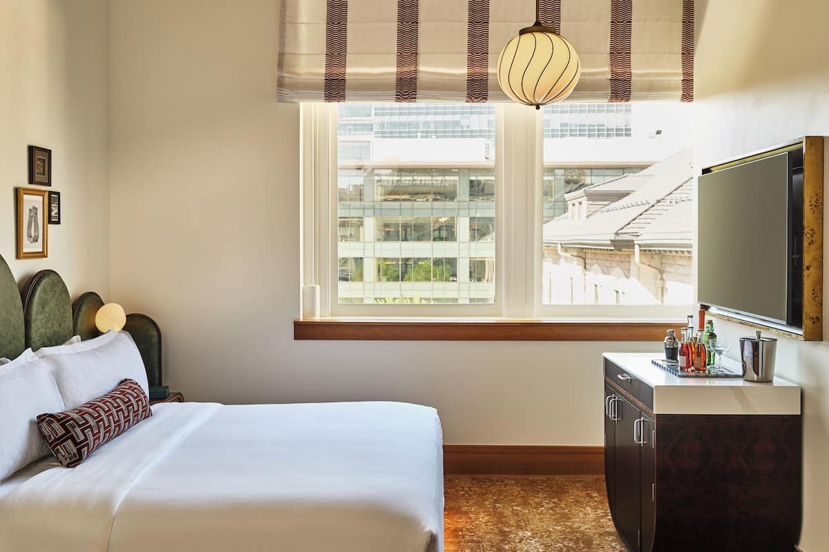 Une chambre à coucher comprenant un lit, une commode, une fenêtre avec des stores et un plafond recouvert d’un papier peint à motifs floraux.