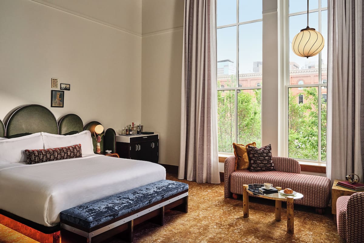 Une chambre d’hôtel avec un grand lit, une banquette, un canapé, une table basse et une vue sur la verdure par la fenêtre.